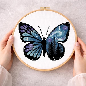 Puede incluir: Bordado de punto de cruz de una mariposa con un dise&ntilde;o de cielo nocturno. La mariposa tiene alas azules y moradas con una escena de noche estrellada en un lado y un bosque en el otro. El bordado est&aacute; en un aro de madera.