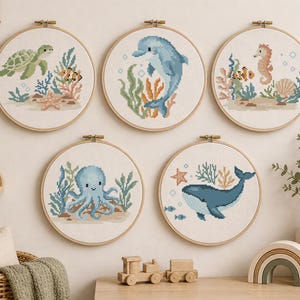 May include: Five round embroidery hoops with cross-stitch designs of sea creatures: a turtle, dolphin, seahorse, octopus, and whale. Each design includes coral, seaweed, and other marine elements, all in a nursery setting.