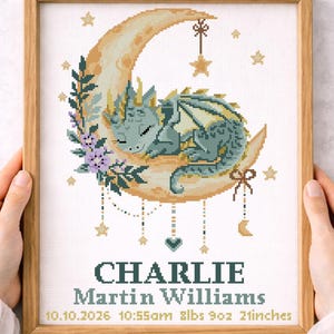 May include: A framed cross-stitch pattern featuring a sleeping dragon on a crescent moon. The design includes stars, floral accents, and the name "CHARLIE" with birth details: 8lbs 9oz, 21 inches. The text "Cross stitch pattern" is also visible.