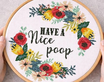 May include: A cross-stitch embroidery hoop with the phrase "Have a Nice Poop" in black text, surrounded by a floral wreath of red, yellow, and teal flowers. The hoop is held by hands.