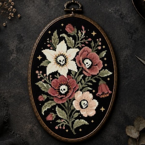 May include: An oval embroidery hoop containing a cross-stitch piece on black fabric. The design features a floral arrangement with white, pink, and red flowers, each with a small skull at its center, surrounded by green leaves and gold stars.