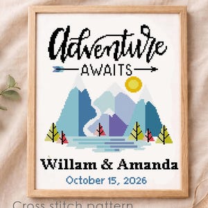 Könnte beinhalten: Ein gerahmtes Kreuzstichdesign mit den Worten "Adventure Awaits" über einer Berglandschaft mit einer Sonne. Darunter steht "Willam & Amanda" und "October 15, 2026". Das Kunstwerk befindet sich in einem Holzrahmen.