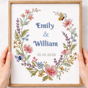Könnte beinhalten: Ein gerahmtes Kreuzstichmuster mit einem floralen Kranz aus rosa, blauen und lila Blumen und Schmetterlingen. Die Namen "Emily & William" und das Datum "10.10.2026" sind mittig platziert. Der Text "Cross stitch pattern Instant download PDF" befindet sich unten.