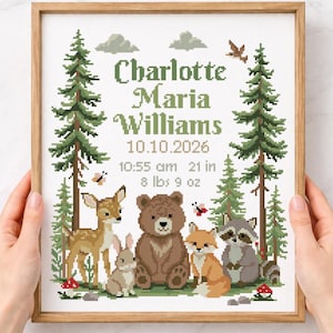 May include: A framed cross-stitch pattern featuring the name Charlotte Maria Williams, the date 10.10.2026, 10:55 am, 21 in, and 8 lbs 9 oz. The design includes green pine trees, a deer, a rabbit, a bear, a fox, a raccoon, butterflies, and mushrooms.