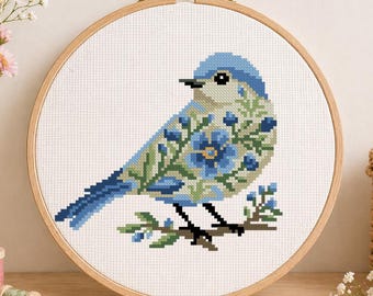 Op de afbeelding: Kruissteekborduurwerk van een blauwe vogel met bloemendetails, weergegeven in een houten hoepel. De vogel zit op een tak met groene bladeren en blauwe bessen. De achtergrond is wit en de hoepel staat op een houten oppervlak.