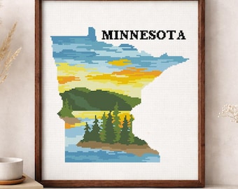 May include: Framed cross-stitch art featuring the outline of Minnesota, filled with a landscape scene of a lake, trees, and a sunset. The word "MINNESOTA" is stitched above the landscape. The artwork is displayed on a white surface.