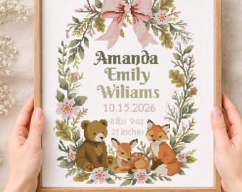 May include: A framed cross-stitch pattern featuring a floral wreath with a pink bow at the top. Inside the wreath are the names Amanda Emily Wiliams, the date 10.15.2026, and the birth details 8 lbs 9 oz and 21 inches, with a bear, deer, and fox.