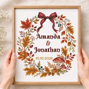 Könnte beinhalten: Ein gerahmtes Kreuzstichmuster mit einem herbstlichen Kranz aus orangefarbenen, roten und braunen Blättern, Beeren und Pilzen. Oben befindet sich eine dunkelrote Schleife. Der Text in der Mitte lautet Amanda & Jonathan und das Datum 10.10.2026.