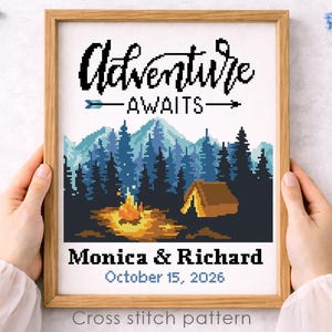以下が含まれることがあります： 「Adventure Awaits」の文字が書かれた額入りのクロスステッチパターン。テントとキャンプファイヤーのある森の風景が描かれています。下には「Monica & Richard, October 15, 2026」と書かれています。フレームは茶色です。