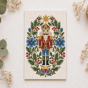 May include: A rectangular cross-stitch embroidery on light fabric featuring a central nutcracker figure in a red and blue uniform, surrounded by an oval wreath of green pine branches, red berries, and blue flowers, topped with a yellow star.