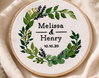 Könnte beinhalten: Ein runder hölzerner Stickrahmen mit weißem Stoff und einem Kreuzstichmotiv. Das Design zeigt einen grünen botanischen Kranz, der den schwarzen Text Melissa & Henry sowie das Datum 10.10.20 in einer einfachen, klaren Schriftart umschließt.