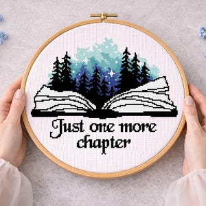 May include: Cross-stitch embroidery in a wooden hoop. The design features an open book with a forest scene and the text "Just one more chapter". The color palette includes white, black, and shades of blue and green.