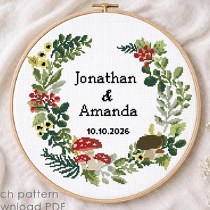 May include: A cross-stitch pattern in a wooden hoop featuring a floral wreath design. The wreath includes green leaves, red berries, and mushrooms. The names "Jonathan & Amanda" and the date "10.10.2026" are centered.