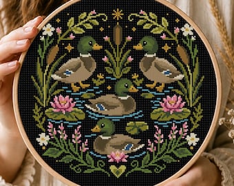 May include: A circular wooden embroidery hoop containing a cross-stitch pattern on black fabric. The design features four mallard ducks swimming in water surrounded by green reeds, cattails, pink water lilies, and small white flowers.