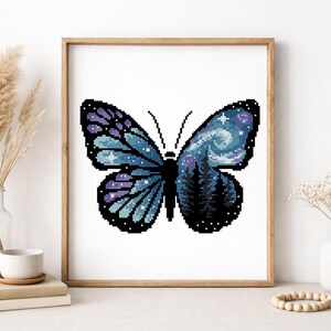 Puede incluir: Una obra de arte en punto de cruz enmarcada que representa una mariposa. Las alas de la mariposa son azules y moradas, con un cielo nocturno estrellado y una escena de bosque. La obra se presenta en un marco de madera.