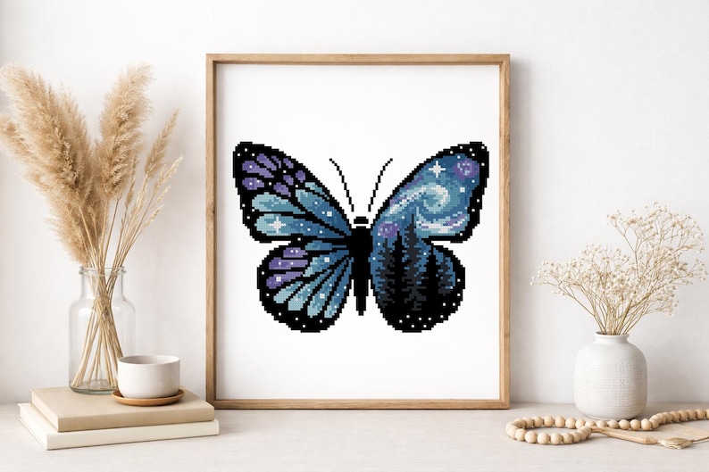 Puede incluir: Una obra de arte en punto de cruz enmarcada que representa una mariposa. Las alas de la mariposa son azules y moradas, con un cielo nocturno estrellado y una escena de bosque. La obra se presenta en un marco de madera.