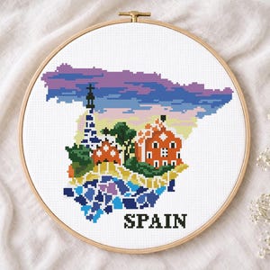 May include: Cross-stitch embroidery of Spain in a wooden hoop. The design features a colorful pixelated map of Spain with iconic buildings and the word "SPAIN" in black block letters. The sky is a gradient of purple and blue.