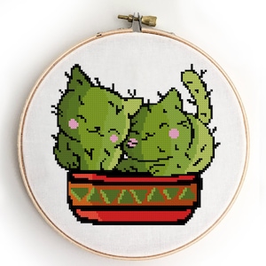 May include: Two green cactus cats in a red and green pot. The cats are hugging and have pink cheeks.