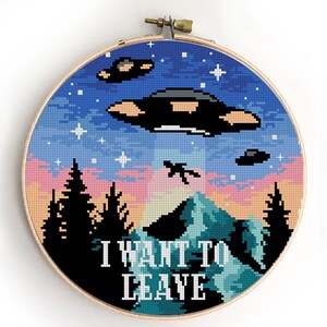 Può includere: Schema di punto croce con un tramonto colorato con montagne, alberi e tre dischi volanti. Il testo "I WANT TO LEAVE" è ricamato sotto la scena.
