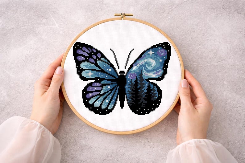 Puede incluir: Bordado de punto de cruz de una mariposa con un dise&ntilde;o de cielo nocturno. La mariposa tiene alas azules y moradas con una escena de noche estrellada en un lado y un bosque en el otro. El bordado est&aacute; en un aro de madera.