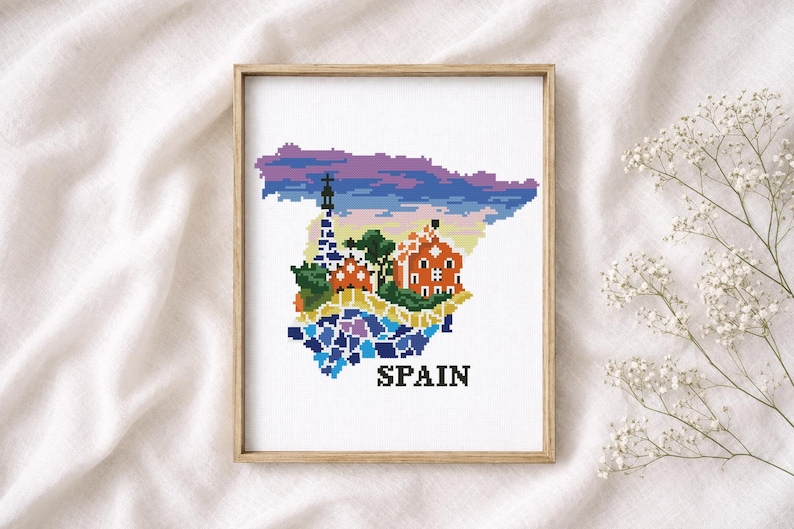 Puede incluir: Arte de punto de cruz del mapa de Espa&ntilde;a, con arquitectura ic&oacute;nica en colores vibrantes. La obra est&aacute; enmarcada en un marco de madera clara e incluye la palabra "SPAIN". El fondo es una tela blanca suave.