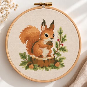 May include: A cross-stitch embroidery project in a round wooden hoop featuring a pixelated orange squirrel sitting on a brown tree stump. The squirrel holds a brown acorn, surrounded by green leaves and small red berries on a light beige fabric background.