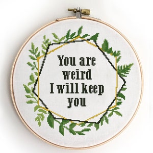 Puede incluir: Un aro de bordado blanco con un diseño floral verde de hojas que rodea un marco geométrico negro y dorado. El texto dentro del marco dice "You are weird I will keep you".