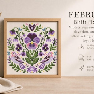 May include: A framed cross-stitch pattern featuring a floral design with violets and birds in shades of purple, green, and pink. The text reads "FEBRUARY Birth Flowers" and describes violets as symbols of humility and devotion.