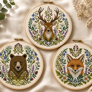 Könnte beinhalten: Drei runde Stickrahmen aus Holz mit Kreuzstichmotiven auf weißem Stoff. Jeder Rahmen zeigt ein zentrales Tierporträt – einen Hirsch, einen Braunbären und einen Fuchs – umgeben von einem detaillierten Kranz aus bunten Wildblumen und grünem Laub.