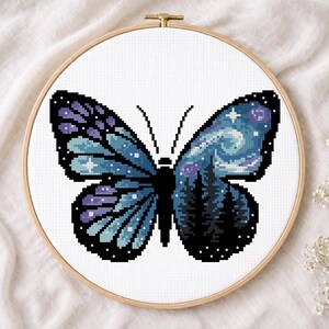 Puede incluir: Bordado de punto de cruz de una mariposa con un dise&ntilde;o de cielo nocturno. La mariposa tiene alas azules y moradas con un patr&oacute;n de estrellas y una silueta de bosque. El bordado est&aacute; en un aro de madera.