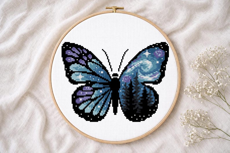 Puede incluir: Bordado de punto de cruz de una mariposa con un dise&ntilde;o de cielo nocturno. La mariposa tiene alas azules y moradas con un patr&oacute;n de estrellas y una silueta de bosque. El bordado est&aacute; en un aro de madera.