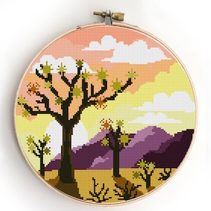 May include: Cross stitch pattern of a desert landscape with a Joshua tree, mountains, and a sunset sky. The pattern is in a circular embroidery hoop.