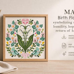 May include: A framed cross-stitch design featuring a floral wreath with pink and white flowers, green leaves, and two blue birds. The text reads "MAY Birth Flowers" symbolizing sweetness, humility, hope, and the return of happiness.