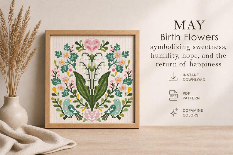 May include: A framed cross-stitch design featuring a floral wreath with pink and white flowers, green leaves, and two blue birds. The text reads "MAY Birth Flowers" symbolizing sweetness, humility, hope, and the return of happiness.
