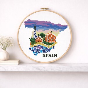 Puede incluir: Arte de bordado a punto de cruz que presenta un mapa pixelado de Espa&ntilde;a con lugares emblem&aacute;ticos. El dise&ntilde;o incluye una iglesia, una torre y la palabra "SPAIN" en negro. La obra de arte se exhibe en una pared blanca sobre una estanter&iacute;a de m&aacute;rmol.