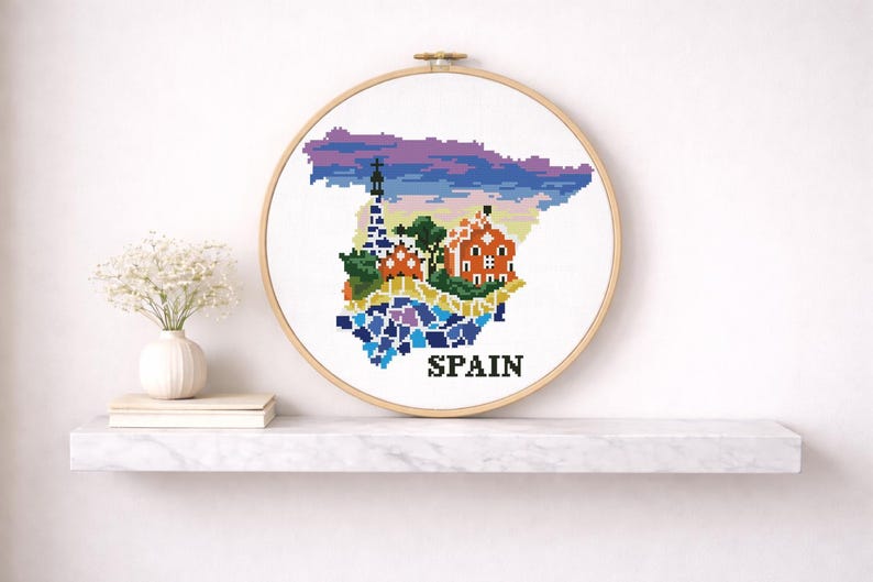 Puede incluir: Arte de bordado a punto de cruz que presenta un mapa pixelado de Espa&ntilde;a con lugares emblem&aacute;ticos. El dise&ntilde;o incluye una iglesia, una torre y la palabra "SPAIN" en negro. La obra de arte se exhibe en una pared blanca sobre una estanter&iacute;a de m&aacute;rmol.