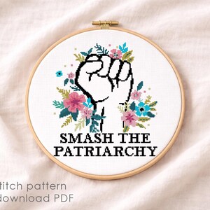 May include: A circular wooden embroidery hoop containing a cross stitch design on white fabric. The design features a black outlined raised fist surrounded by pink and blue flowers with green leaves, and the text SMASH THE PATRIARCHY in black.