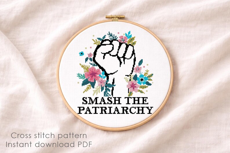 May include: A circular wooden embroidery hoop containing a cross stitch design on white fabric. The design features a black outlined raised fist surrounded by pink and blue flowers with green leaves, and the text SMASH THE PATRIARCHY in black.