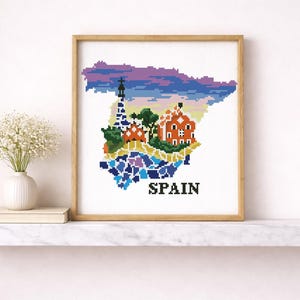 Puede incluir: Una obra de arte en punto de cruz enmarcada que representa el mapa de Espa&ntilde;a, con ilustraciones pixeladas coloridas de monumentos y un cielo vibrante. La palabra "SPAIN" est&aacute; bordada en negro en la parte inferior. La obra se exhibe en una estanter&iacute;a blanca.