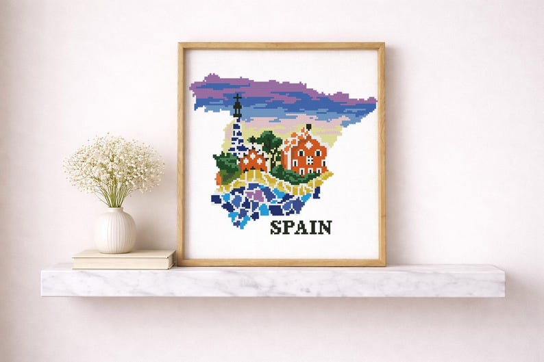 Puede incluir: Una obra de arte en punto de cruz enmarcada que representa el mapa de Espa&ntilde;a, con ilustraciones pixeladas coloridas de monumentos y un cielo vibrante. La palabra "SPAIN" est&aacute; bordada en negro en la parte inferior. La obra se exhibe en una estanter&iacute;a blanca.