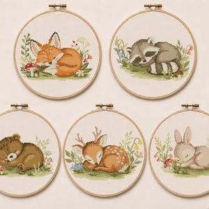 Könnte beinhalten: Fünf Kreuzstich-Stickrahmen mit schlafenden Waldtieren: ein Fuchs, ein Waschbär, ein Bär, ein Reh und ein Kaninchen. Jedes Design enthält Blumen- und Pilz-Akzente, gesetzt auf cremefarbenem Stoff in einem Holzrahmen.