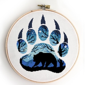 Puede incluir: Un patrón de punto de cruz de una huella de pata de oso con una silueta de un oso dentro. La huella de la pata es azul y blanca con un fondo de cielo estrellado.