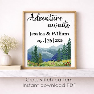 May include: A framed cross-stitch pattern with the words "Adventure awaits" and the names Jessica & Wiliam, with the date September 26, 2026. The artwork depicts a mountain landscape with trees and flowers. The frame is a light brown color.