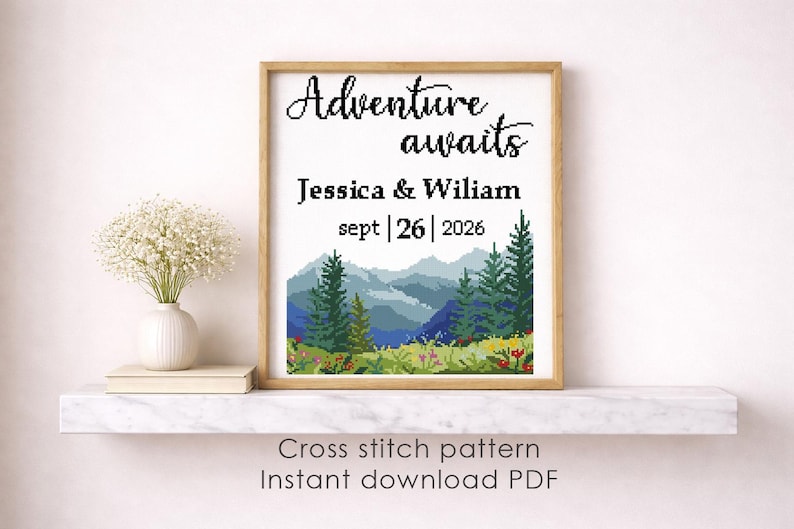 May include: A framed cross-stitch pattern with the words "Adventure awaits" and the names Jessica & Wiliam, with the date September 26, 2026. The artwork depicts a mountain landscape with trees and flowers. The frame is a light brown color.