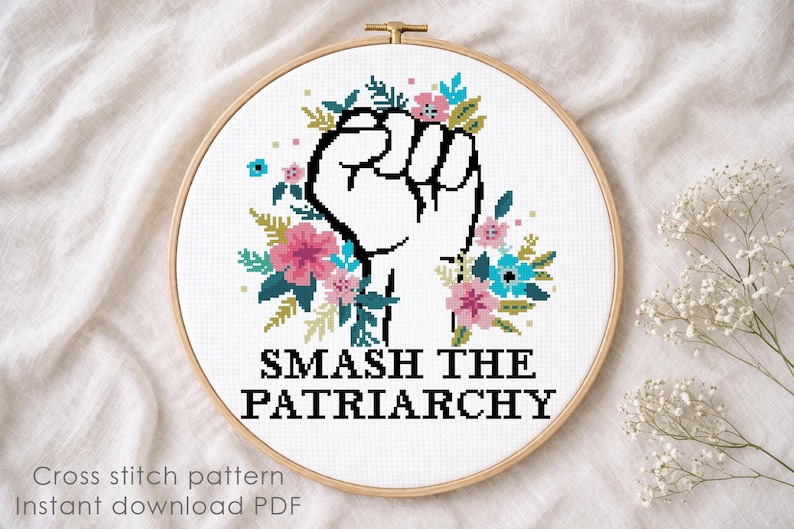 May include: A cross-stitch pattern in a wooden embroidery hoop featuring a raised fist outlined in black, surrounded by pink and blue flowers and green leaves. Below the design, the text reads SMASH THE PATRIARCHY in bold black capital letters.