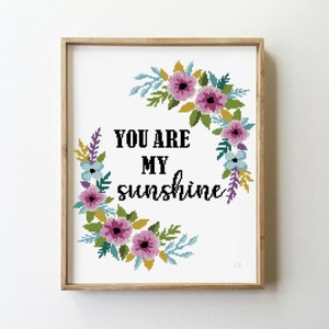 Puede incluir: Patrón de punto de cruz con una corona floral de flores rosas, azules y amarillas. El texto "You are my sunshine" está en el centro de la corona.