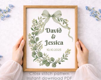 May include: A framed cross-stitch pattern featuring a green wreath with a bow, surrounding the names "David & Jessica" and the date "10.10.2025". The frame is light brown. The text "Cross stitch pattern" and "Instant download PDF" are at the bottom.
