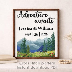 May include: A framed cross-stitch pattern with the words "Adventure awaits" above the names Jessica & Wiliam and the date Sept 26, 2026. The artwork depicts a mountain landscape with trees and flowers. The frame is brown.