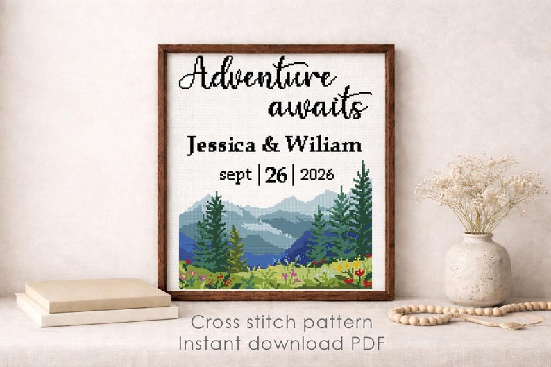 May include: A framed cross-stitch pattern with the words "Adventure awaits" above the names Jessica & Wiliam and the date Sept 26, 2026. The artwork depicts a mountain landscape with trees and flowers. The frame is brown.