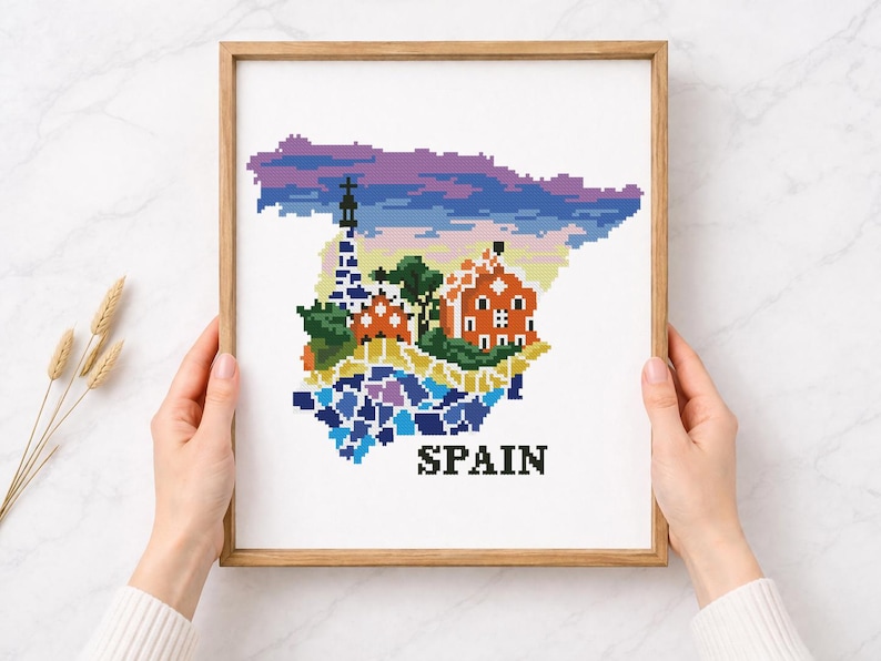 Puede incluir: Una obra de arte en punto de cruz enmarcada que representa el mapa de Espa&ntilde;a, con ilustraciones pixeladas de monumentos y un cielo colorido. La palabra "SPAIN" est&aacute; bordada en letras negras en la parte inferior.